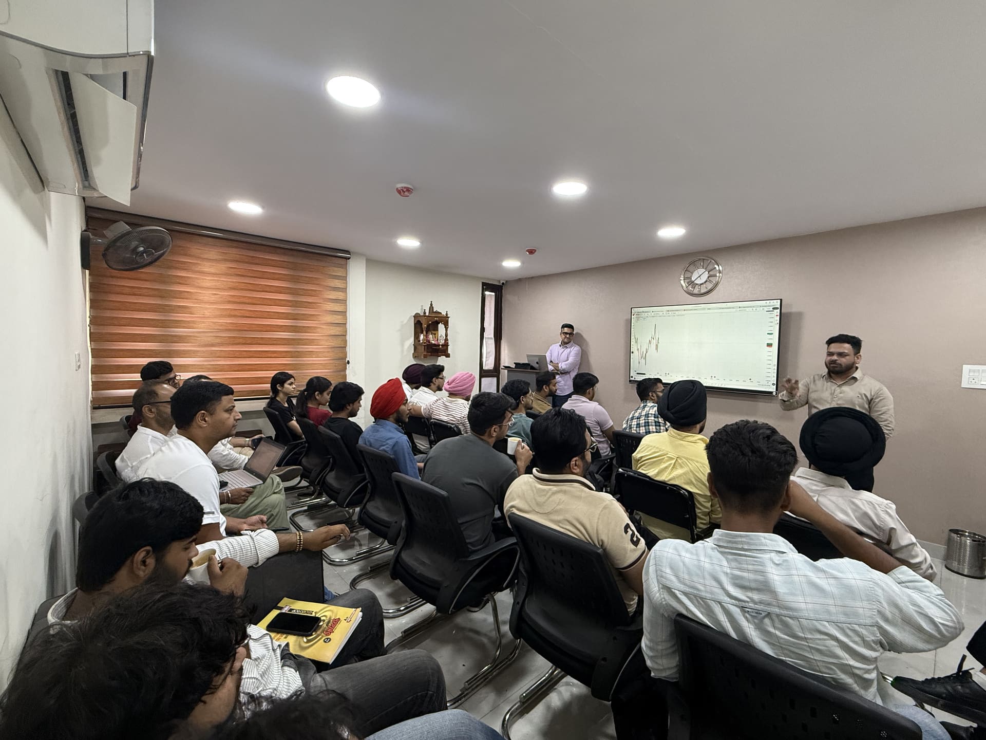 Mentors teaching a full classroom batch at Wise Bucks, Chandigarh, with a candlestick chart on the screen